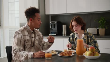 A-young-adult-girl-with-brown-hair-and-a-bob-hairstyle-in-a-green-checkered-shirt-communicates-with-her-young-boyfriend-with-Black-brunette-skin-color-at-a-black-kitchen-table-in-the-kitchen-in-a-modern-apartment