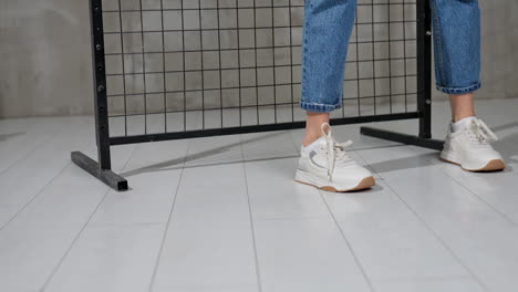 girl turns around demonstrating the comfortable white sneakers. female in blue jeans walks in stylish footwear. black grate at backdrop.