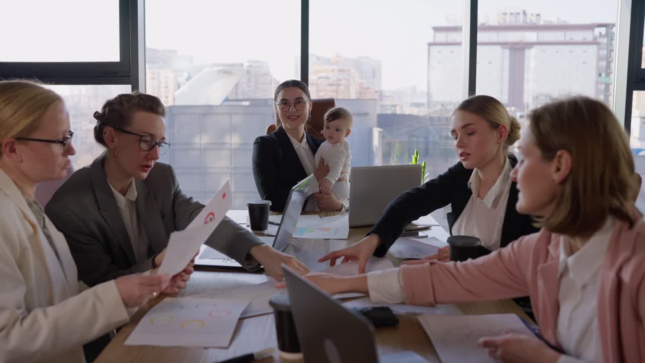 retrato de una chica morena confiada con gafas redondas en un traje de negocios que se sienta en una mesa en la oficina y sostiene a un niño pequeño en sus brazos durante una reunión y pensando en su idea y plan en una oficina con grandes ventanas en la mesa