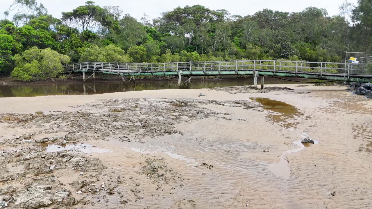 Drone glides over sandy estuary and wooden bridge, revealing lush greenery and calm water reflections
