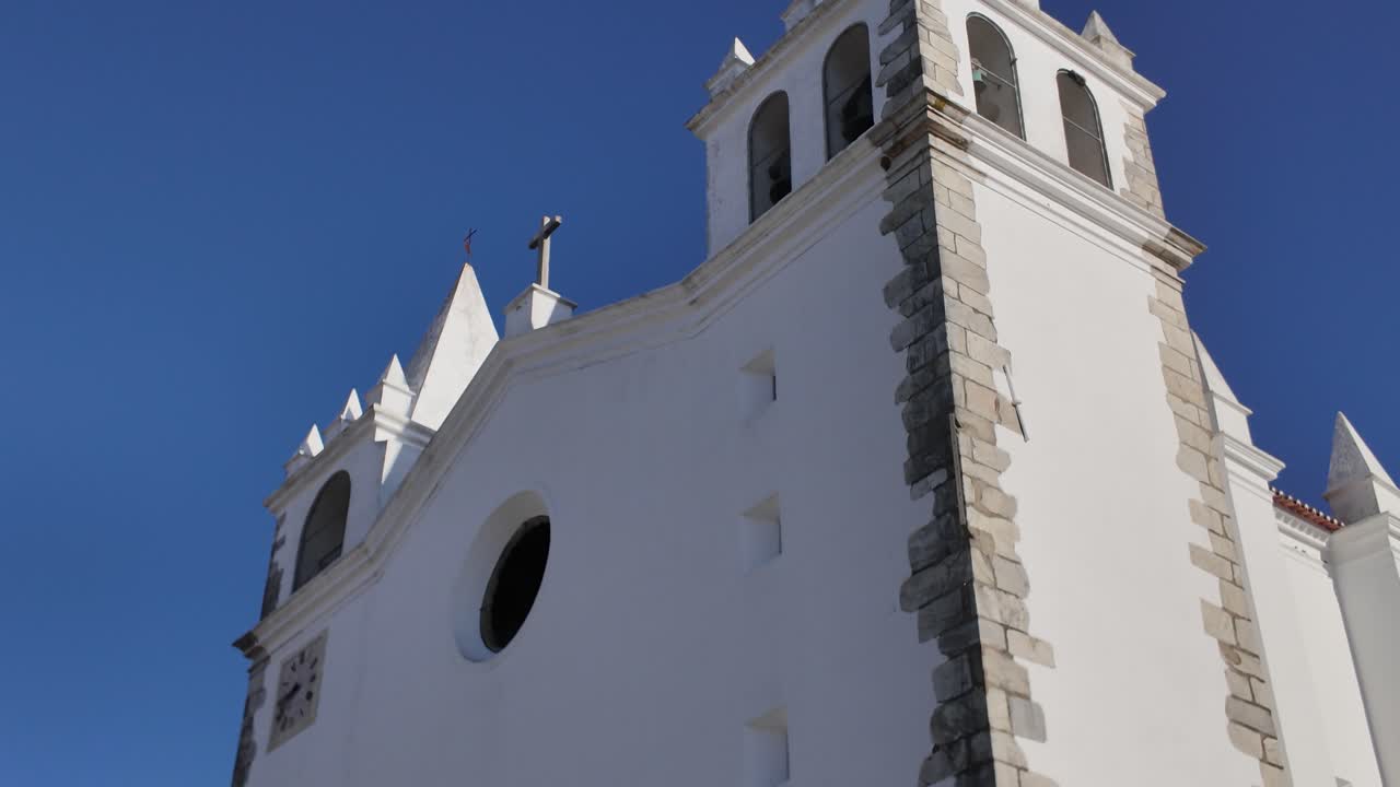 White facade of the Church of Vila Nova da Baronia in Portugal, featuring architectural details and a cross against a clear blue sky