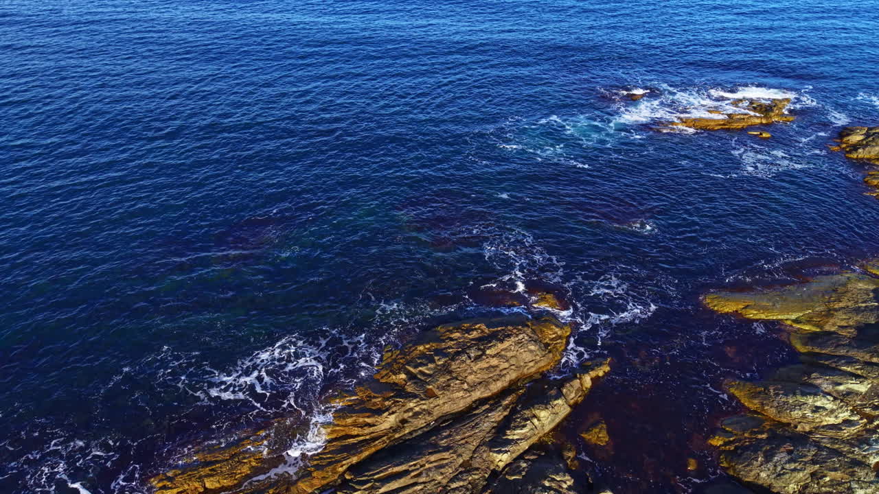 Onde oceaniche vivaci si infrangono contro aspre scogliere sotto un cielo azzurro al tramonto, creando uno spettacolo naturale mozzafiato lungo la costa.