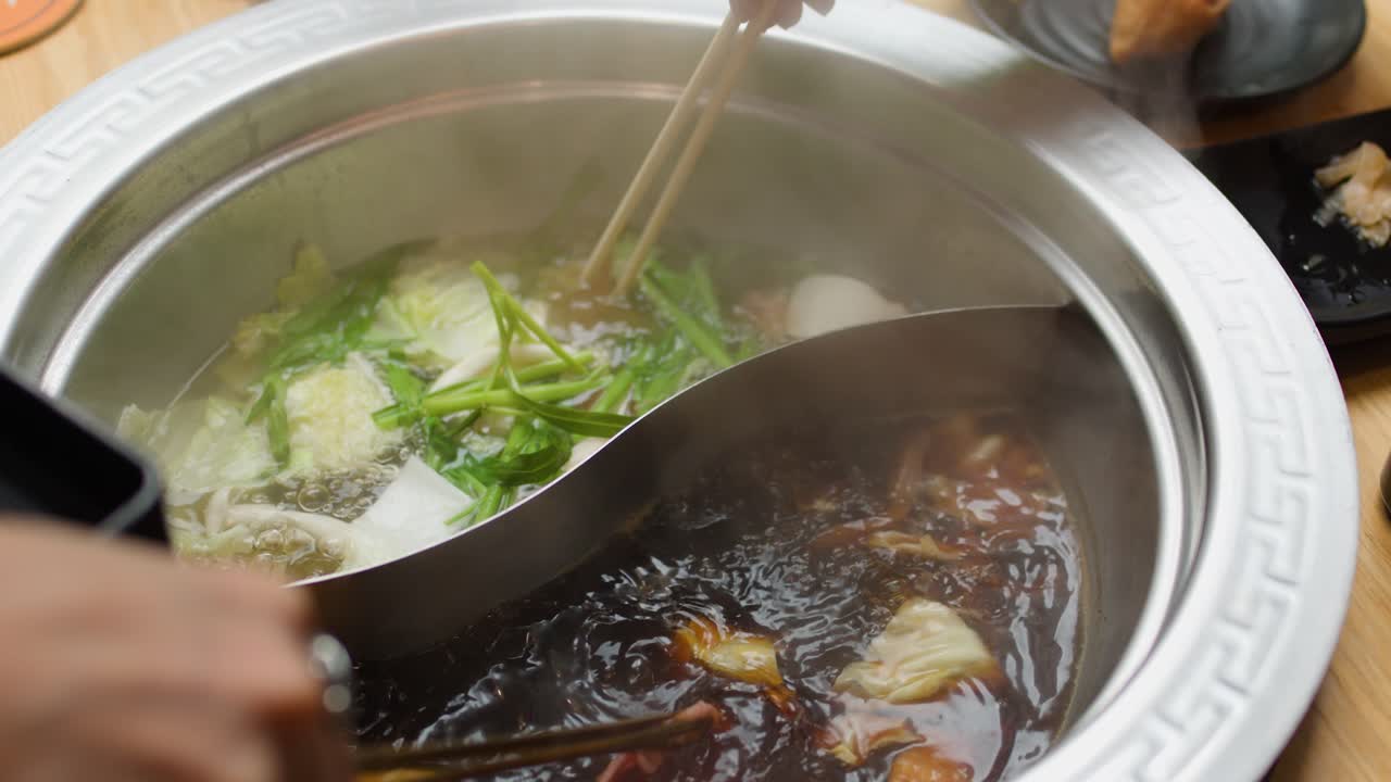 Hand cooks sliced meat in steaming dual broth hotpot with vegetables, natural lighting, close-up view