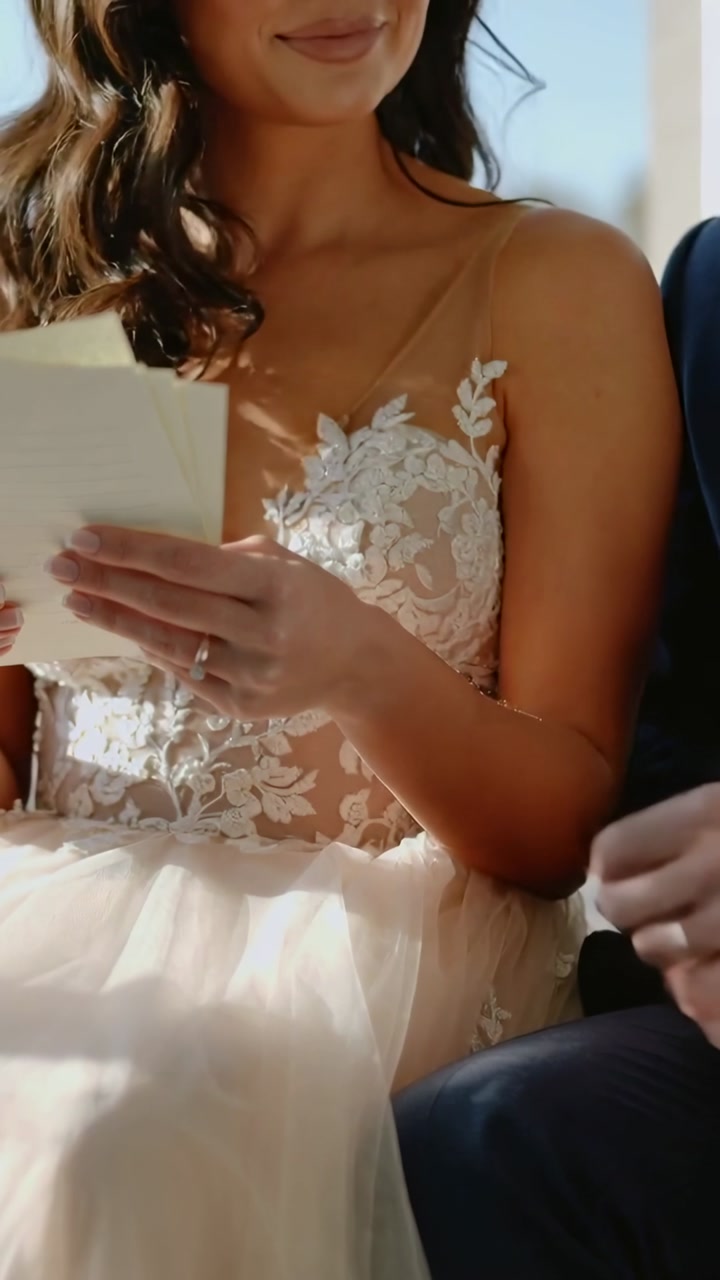 Vertical video: Holding notes, bride reading vows in lace gown at bench, groom touching ring