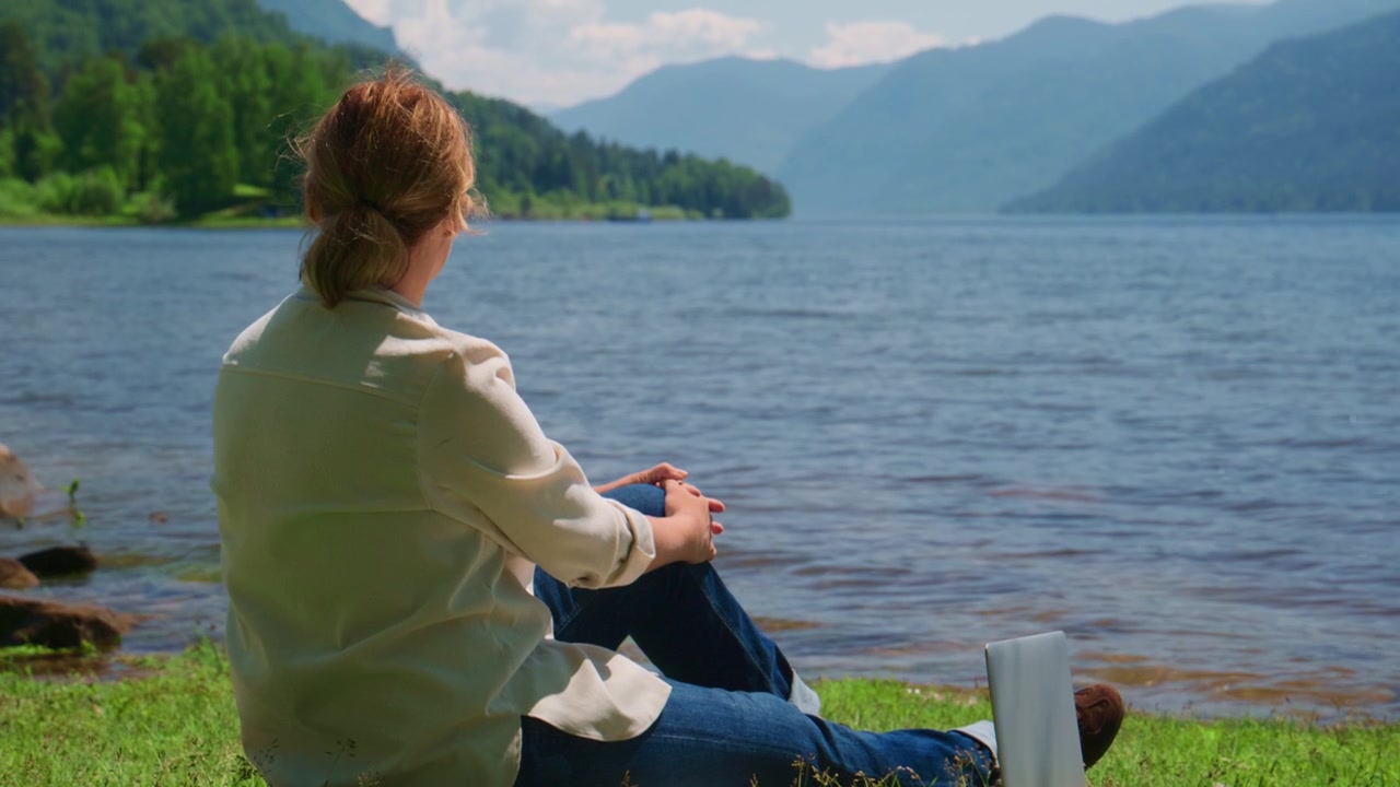 Nainen istuu rauhallisesti tyynen järven rannalla, katse kohti kaukaisia vuoria. Hän vaikuttaa rentoutuneelta nauttien hiljaisesta yksinäisyydestä ulkona. Kohtaus sopii ihanteellisesti matkailuun, meditaatioon tai luontoaiheisiin projekteihin.