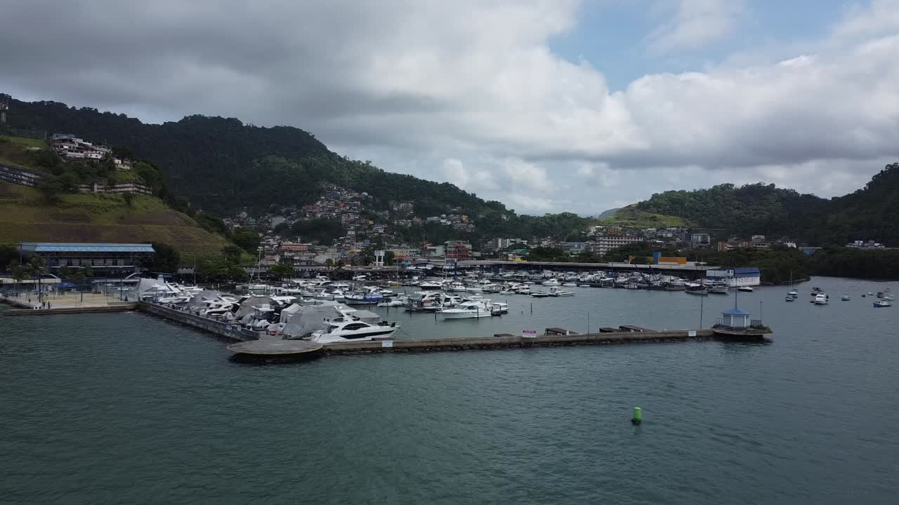 Angra dos Reis marinasına doğru yavaşça yaklaşan, eğlence amaçlı motorlu teknelerin bulunduğu, 4K düşük irtifa drone çekimi. Şehir, dağlar ve Atlantik ormanı arka planda, kısmen bulutlu.