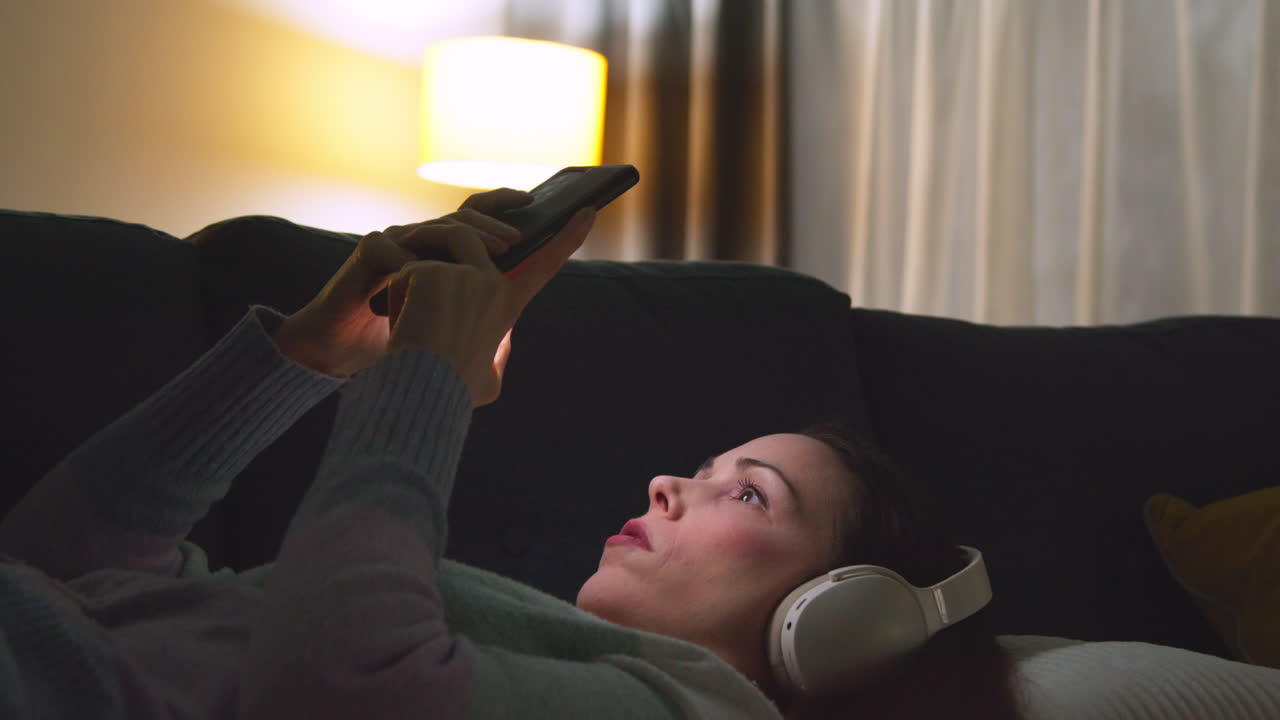 Woman Wearing Wireless Headphones Lying On Sofa At Home At Night Streaming Or Looking At Online Content On Mobile Phone 1