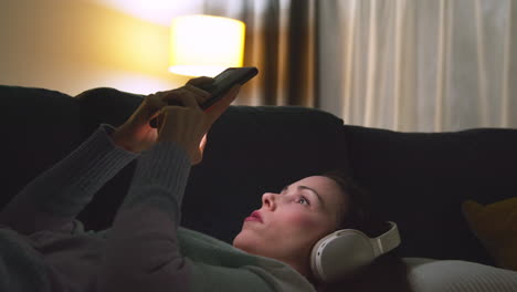 Mujer-Con-Auriculares-Inalámbricos-Recostada-En-Un-Sofá-En-Casa-Por-La-Noche-Transmitiendo-O-Mirando-Contenido-En-Línea-En-Un-Teléfono-Móvil-1