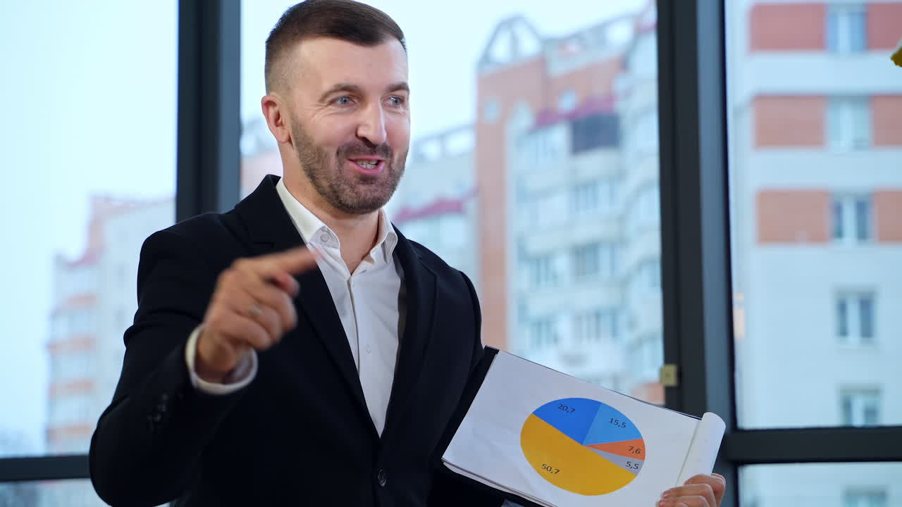 Good-looking business chief talking to his subordinates. Man holding chart and pointing at it with a pen. Blurred background.