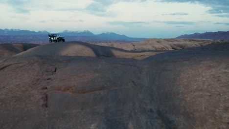 Eine-4K-Drohnenaufnahme-Eines-Hochgefahrenen-Jeeps,-Der-Im-Gelände-Durch-Die-Extreme-Und-Felsige-Wüstenlandschaft-In-Der-Nähe-Von-Moab,-Utah,-Fährt,-Mit-Den-Schneebedeckten-Rocky-Mountains-In-Der-Ferne