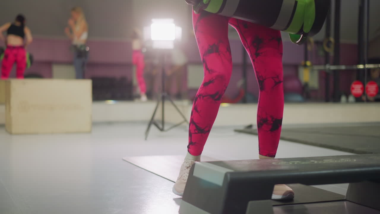 En treningsutøver utfører sit-ups med vekter på matte med step-plattform og studiolys, der sterke rosa treningstights og en sandsekk legger til intensitet, med en trener og en klasse i bakgrunnen, alt i et energisk tempo og med et fokusert uttrykk.