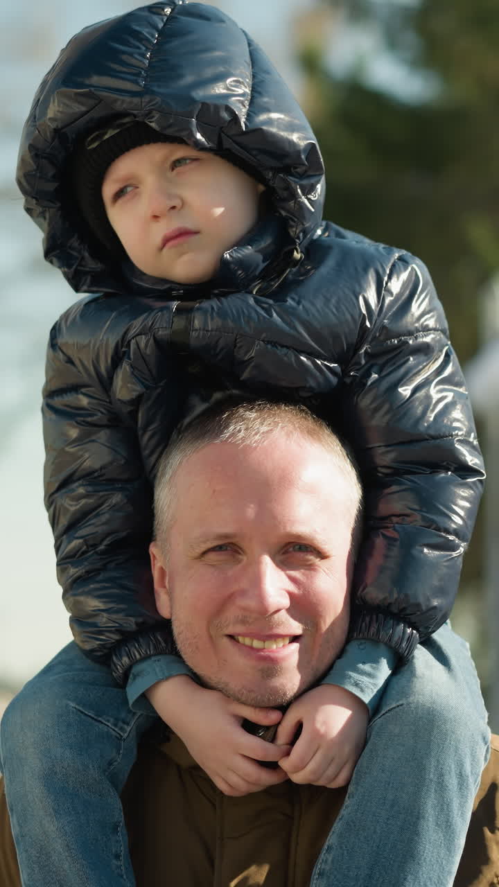 un primer plano de un padre llevando a su hijo en sus hombros sonriendo mientras el niño parece infeliz, con árboles y postes eléctricos detrás de ellos