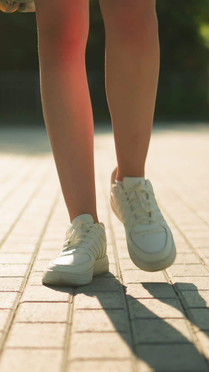 Leg view of individual wearing white canvas shoes walking along interlocked path with shadow cast on ground, with partial view of roller skate, blurred background featuring greenery