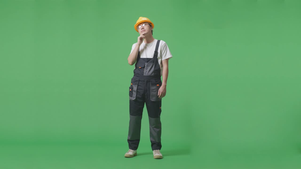 안경과 안전 헬을 착용 한 아시아 남성 노동자가 생각하고 주위를 둘러보고 녹색 스크린 배경 스튜디오에 서있는 동안 지표손가락을 들어 올립니다.
