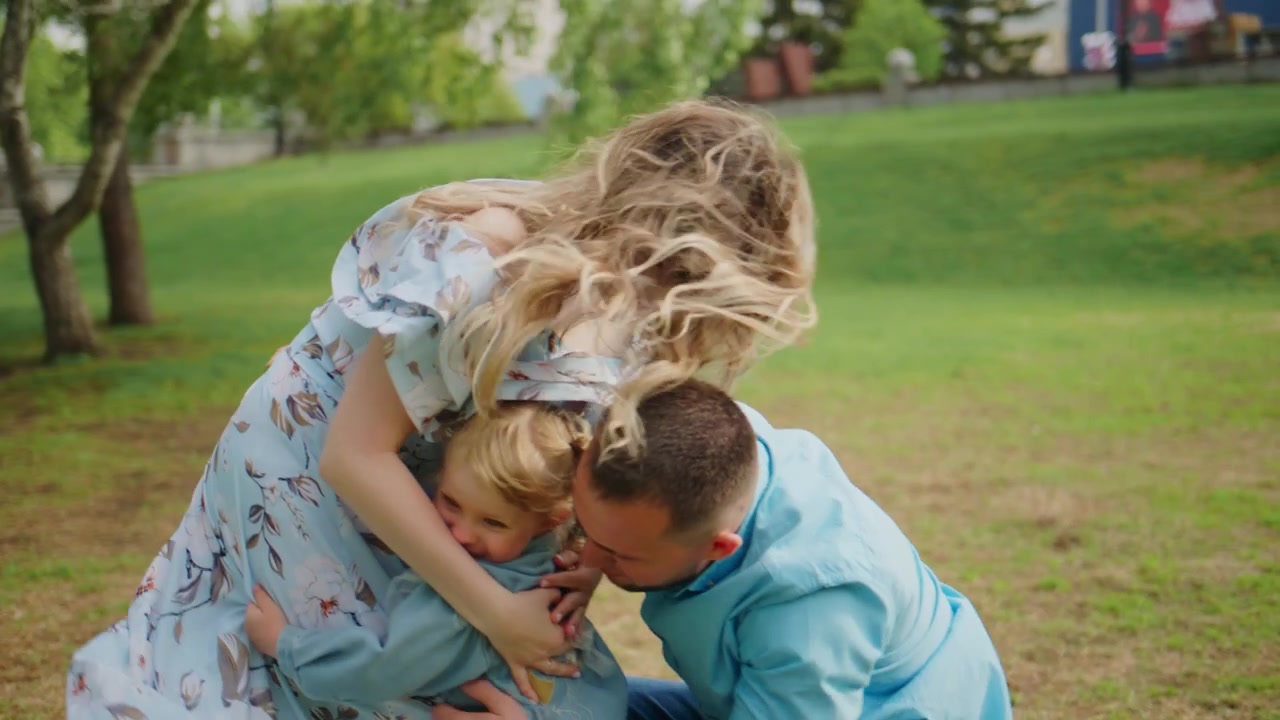 Famiglia che gioca insieme sull'erba assolata. La madre tiene in braccio il bambino mentre il padre si inginocchia. Ridono e sorridono. La scena mostra una famiglia felice che lega all'aperto. Ideale per contenuti su genitorialità, famiglia o stile di vita.