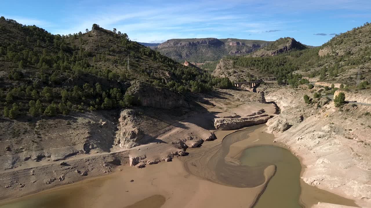 Aerial view of the drought-stricken Fuensanta Reservoir in Yeste, Albacete, featuring a mountainous landscape surrounded by pine trees, dry terrain, and a bridge in the background