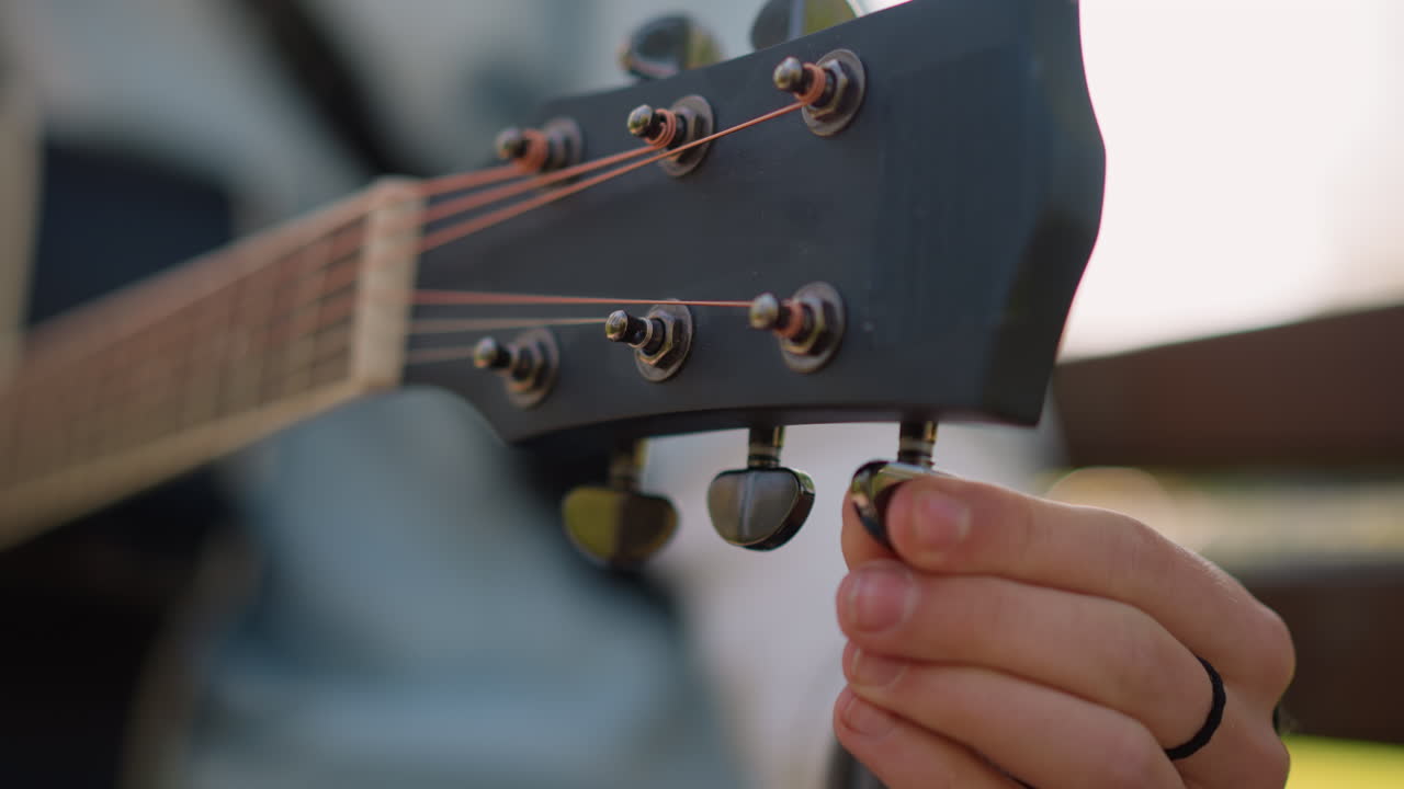 Guitarist tuning acoustic guitar headstock outdoors, hand adjusting tuning pegs, warm copper strings, wood grain, denim shirt, slow delicate motion, focused practice atmosphere, intimate musical craft