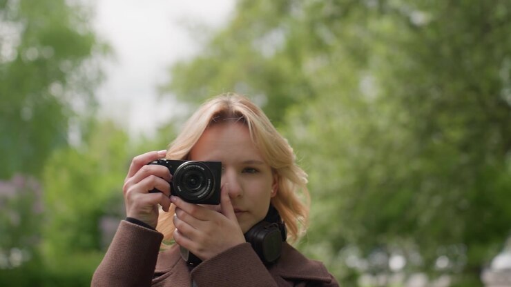 Lady with blonde hair wears brown coat and takes photos using black digital camera while standing outdoors. background features green trees suggesting scene is set in park or garden