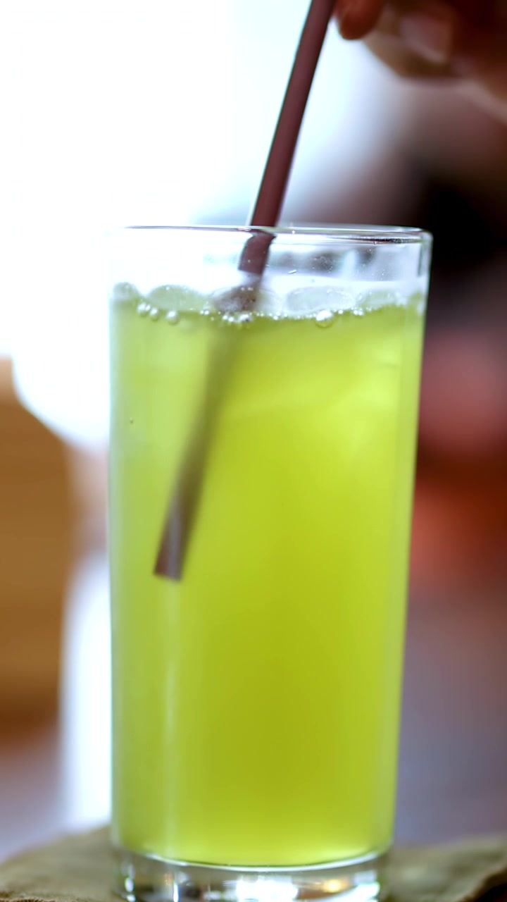 Una mano mescola tè verde freddo con una cannuccia in un luminoso ambiente interno di un ristorante a Bangkok, con messa a fuoco selettiva.