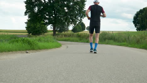 Tiefaufnahme-Der-Schuhe-Und-Beine-Eines-Mannes,-Der-Auf-Einer-Asphaltstraße-Zwischen-Feldern-Läuft