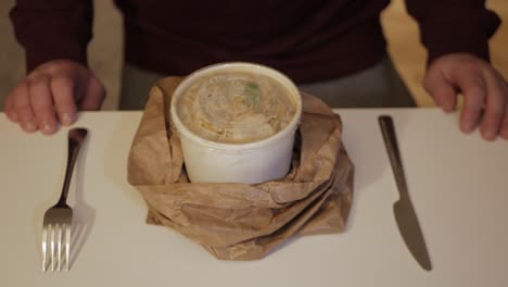un hombre abriendo una bolsa de papel arrugada con un tazón de fideos de curry verde adentro - primer plano