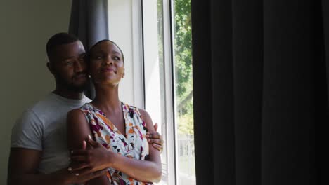Afroamerikanisches-Paar-Umarmt-Sich,-Während-Es-Zu-Hause-Aus-Dem-Fenster-Schaut
