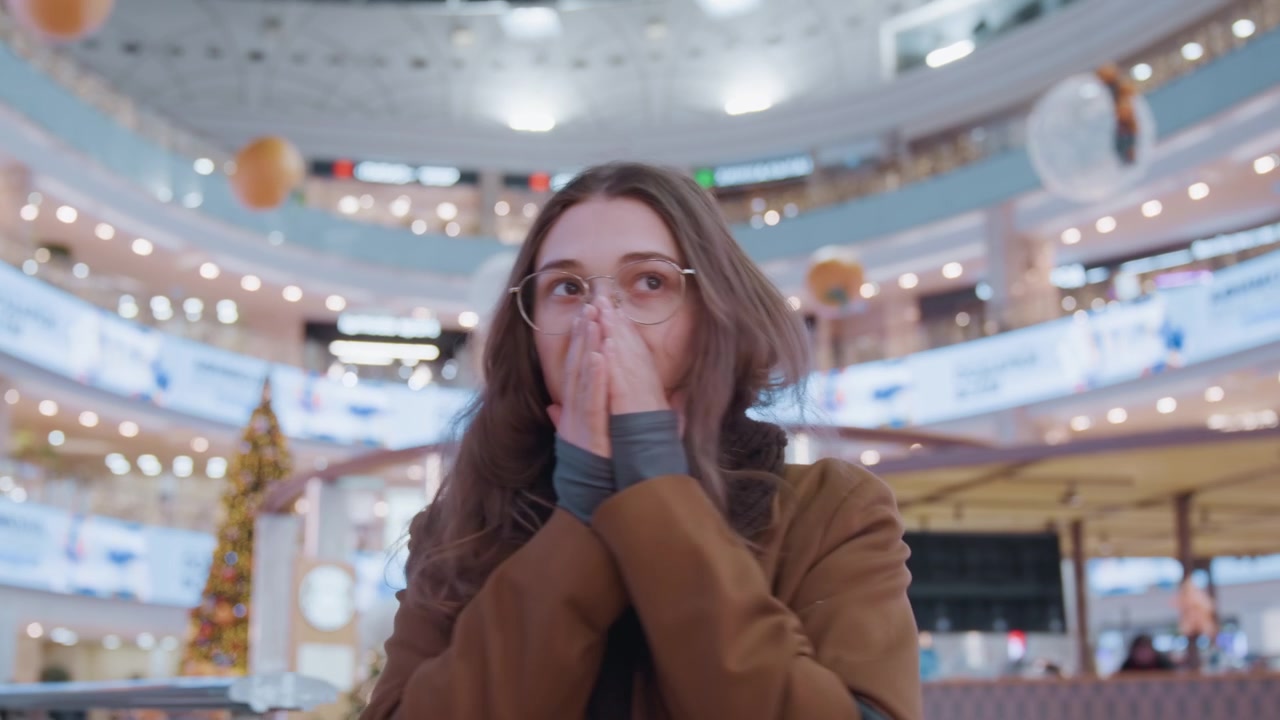 Donna caucasica che osserva l'atrio del centro commerciale, inquadratura ampia degli interni con balconi a spirale, striscioni e insegne LED, acquirenti che si muovono sotto, espressione contemplativa, sciarpa e cappotto, illuminazione morbida e fredda