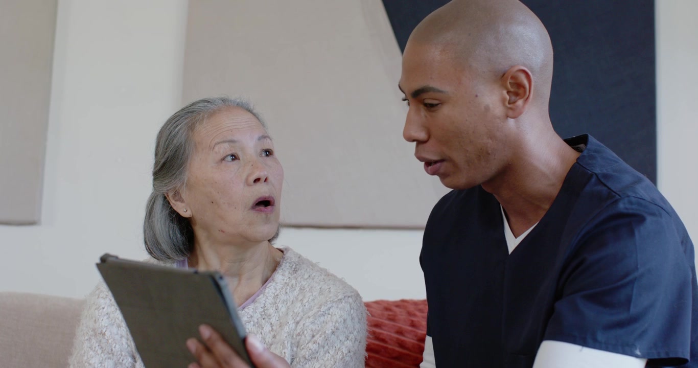 Caregiver diversificato in camice che gira un tablet verso una donna anziana asiatica sul divano, guidando l'interazione