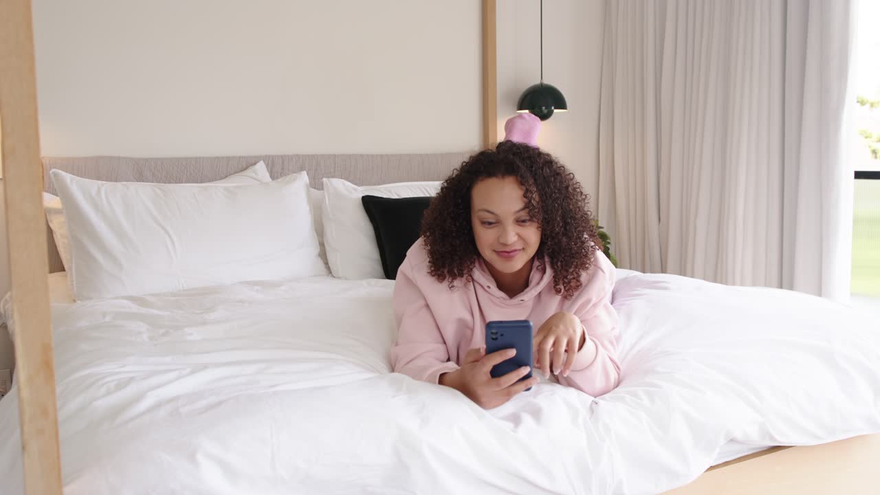 African American woman lying on stomach on bed wearing hoodie, reading on blue phone and tapping
