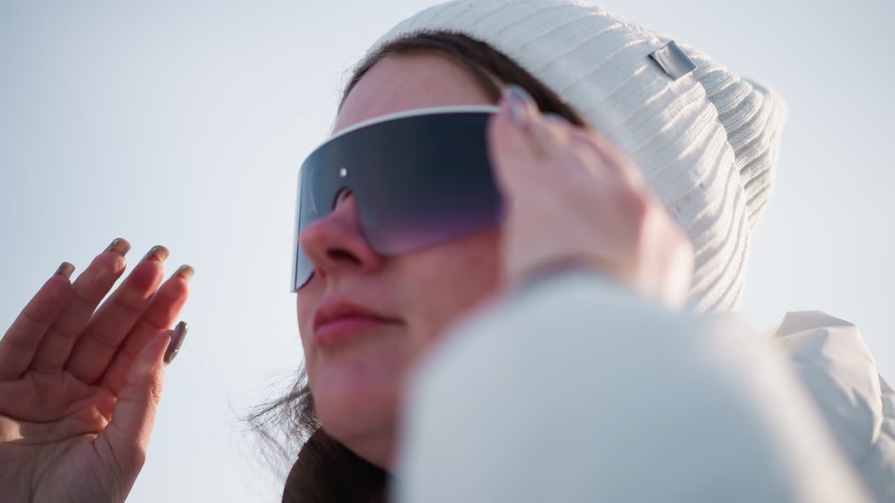 Young artist puts on colored goggles while wearing beanie, hair blowing gently in wind under bright winter sunshine, tinted visor reflecting snowy scene as she adjusts goggles with manicured nails
