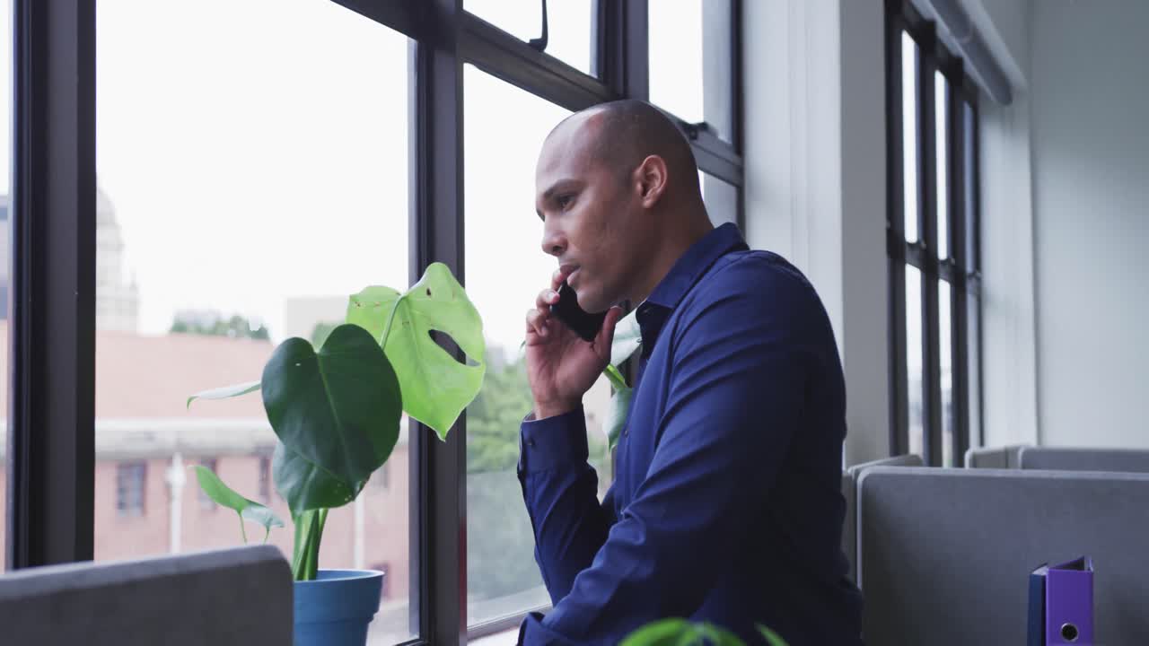 Uomo d'affari di etnia mista in piedi che parla allo smartphone guardando fuori dalla finestra in ufficio.