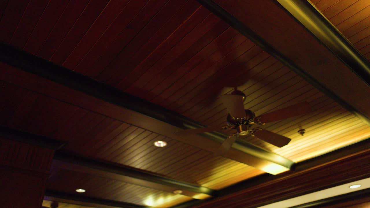 Ceiling fan rotates on wooden ceiling with warm lighting, tropical interior, steady camera angle