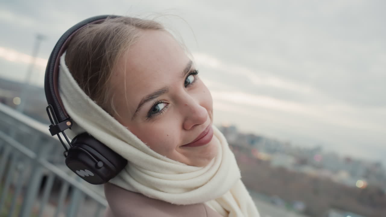 Donna bianca su un tetto con vista sulla città che ascolta le cuffie, con sciarpa e cappotto, che domina lo skyline in una soffusa luce invernale, passeggiata contemplativa, dettagli di moda urbana, punto di osservazione dalla ringhiera, atmosfera panoramica.