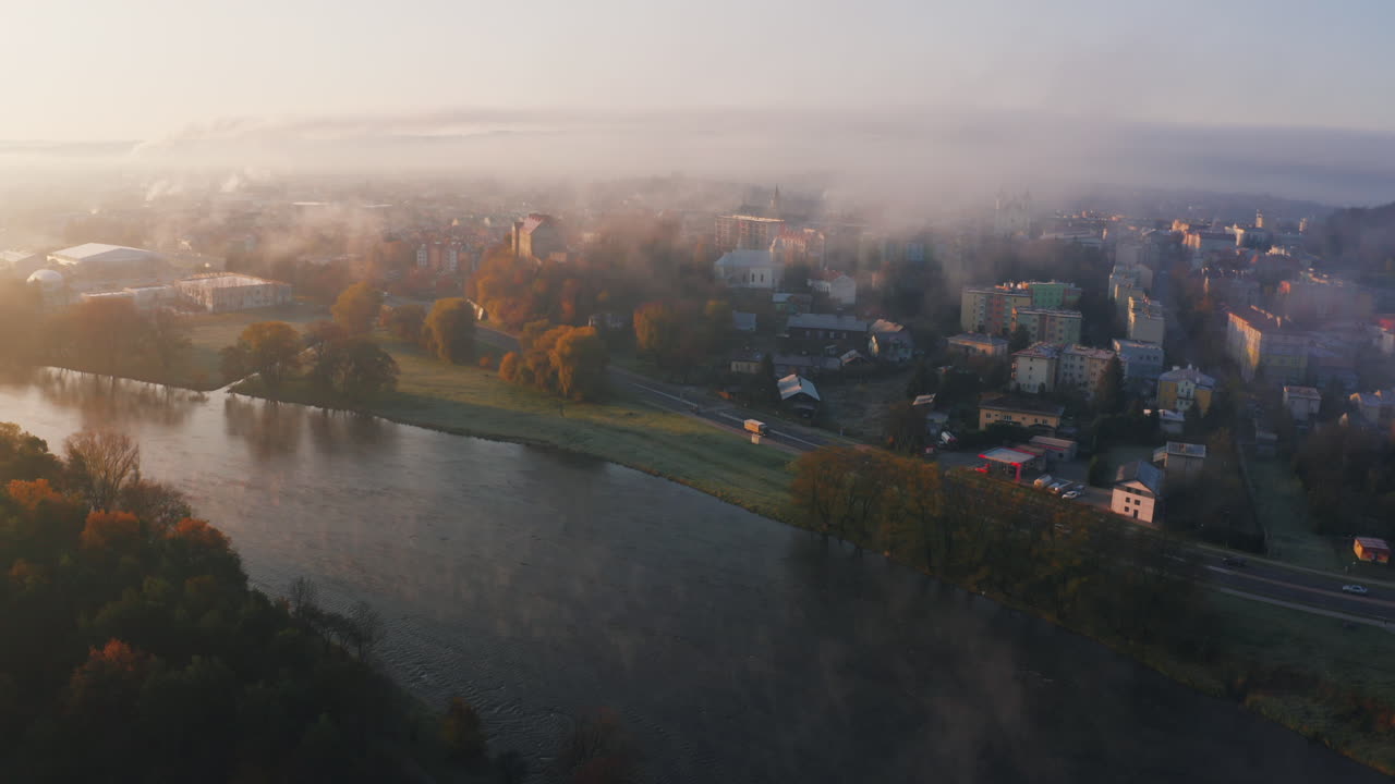 An aerial view of a misty town at sunrise, fog hovering over buildings and a river winding through the landscape in the foreground, creating a vivid, unique, autumn morning scene. Sanok, Poland