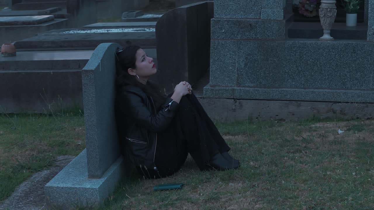 Una ragazza siede appoggiata a una lapide, profondamente assorta in un cimitero di Melbourne durante il tramonto. L'atmosfera malinconica è catturata da una luce soffusa, evidenziando l'ambiente sereno.