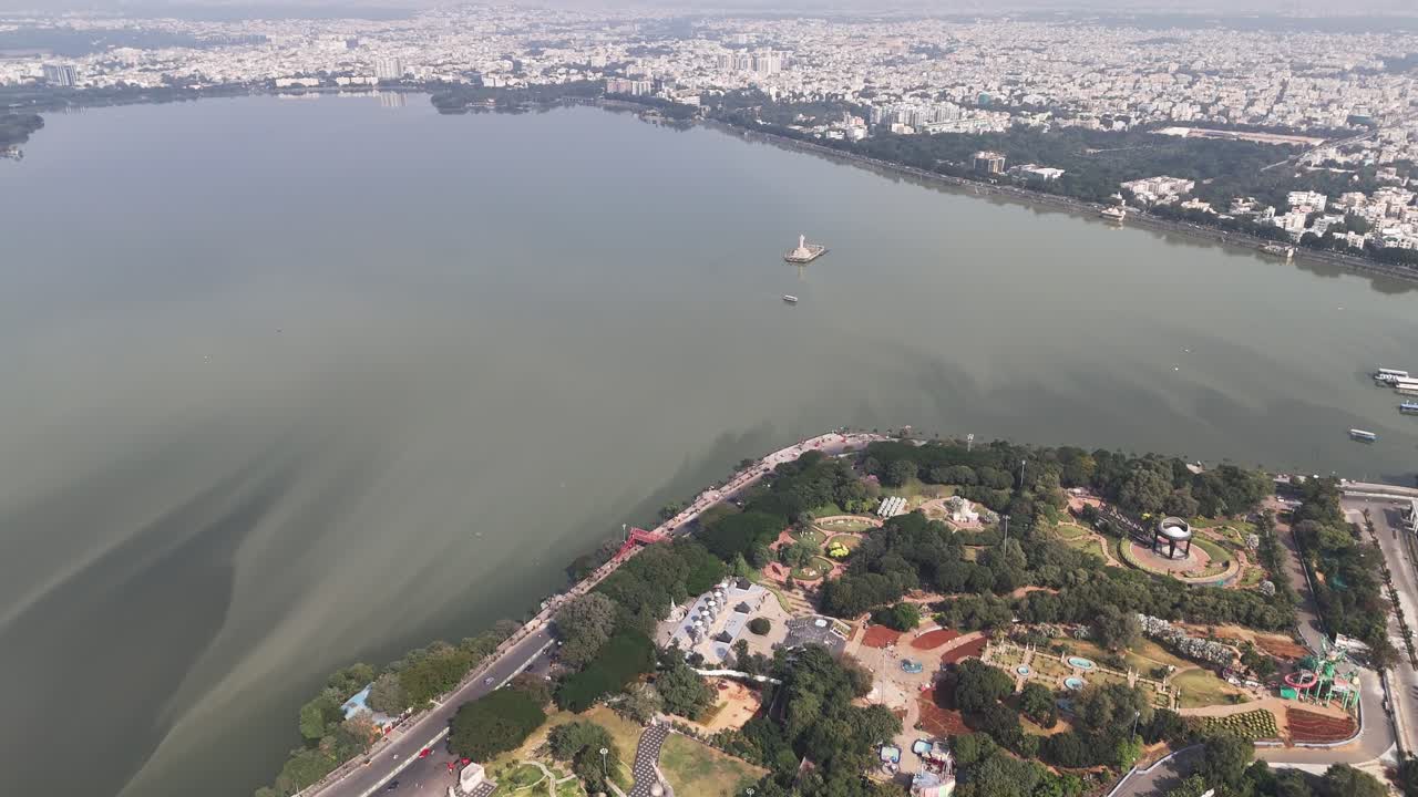 Ripresa video con drone che cattura Hussain Sagar con l'iconica statua del Buddha e una scia di barche che forma motivi circolari sull'acqua serena.