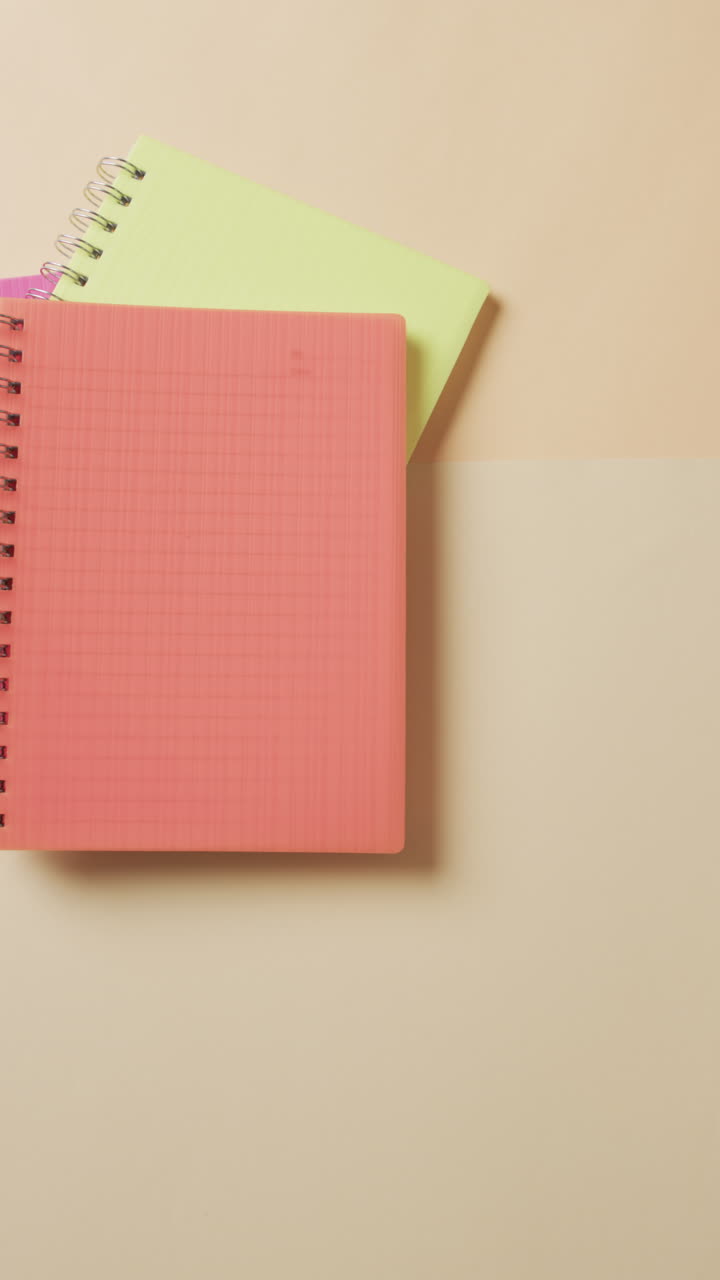 Vertical video of overhead view of colourful notebooks on beige background, in slow motion