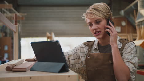 female apprentice carpenter with digital tablet working in furniture workshop making phone call