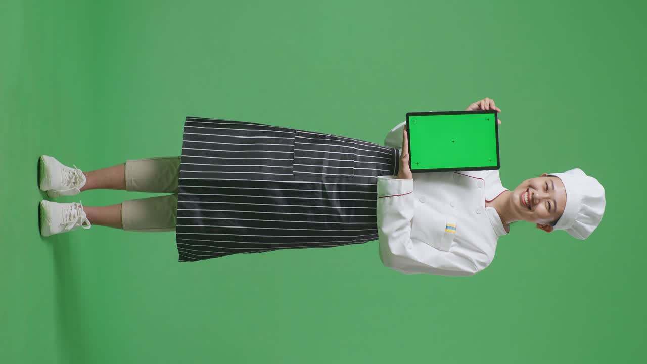 Full Body Of Asian Woman Chef Smiling And Showing Green Screen Tablet To The Camera While Standing In The Green Screen Background Studio
