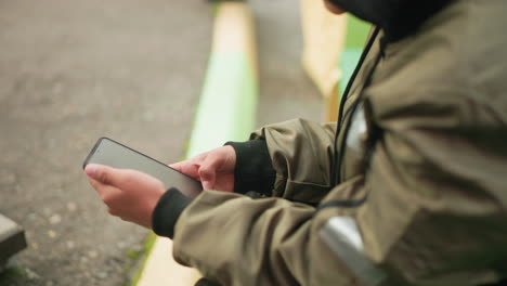 close up side view of kid in jacket holding smartphone with both hands while sitting outdoors, fully engaged in device operation with subtle movement visible in background