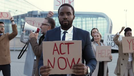 porträt eines afroamerikaners, der act now hält"schild bei einem protest mit multiethnischen geschäftskollegen auf der straße"