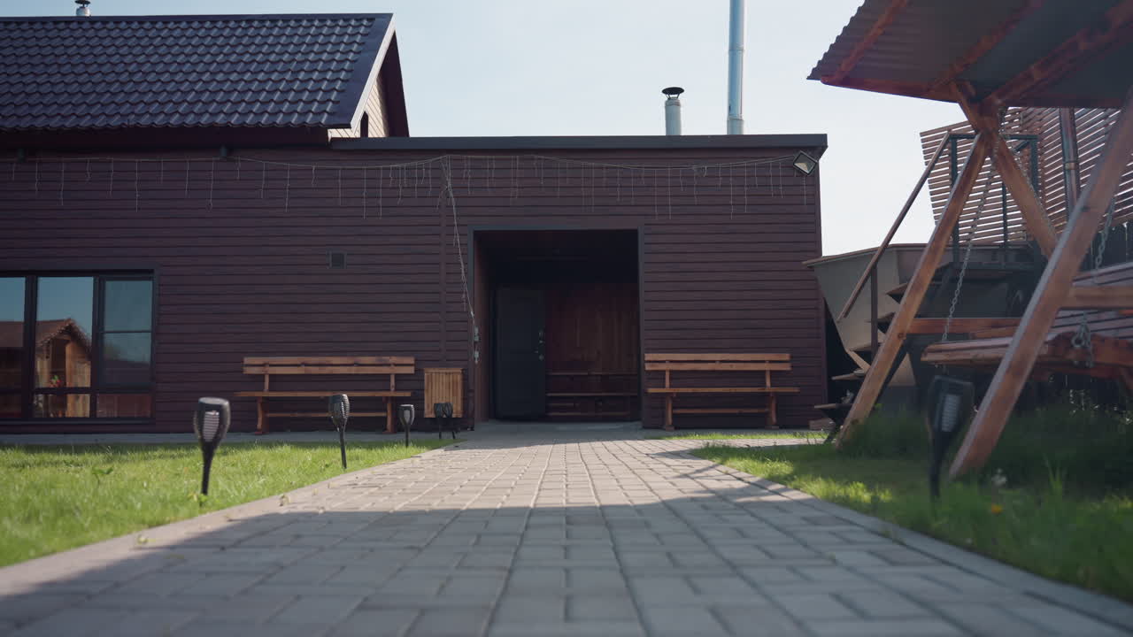 Brown wooden house in sunny fenced compound with small wooden cabin, paved walkway, bench seating, decorative hanging lights, wooden swing, lush grass gently swaying in breeze