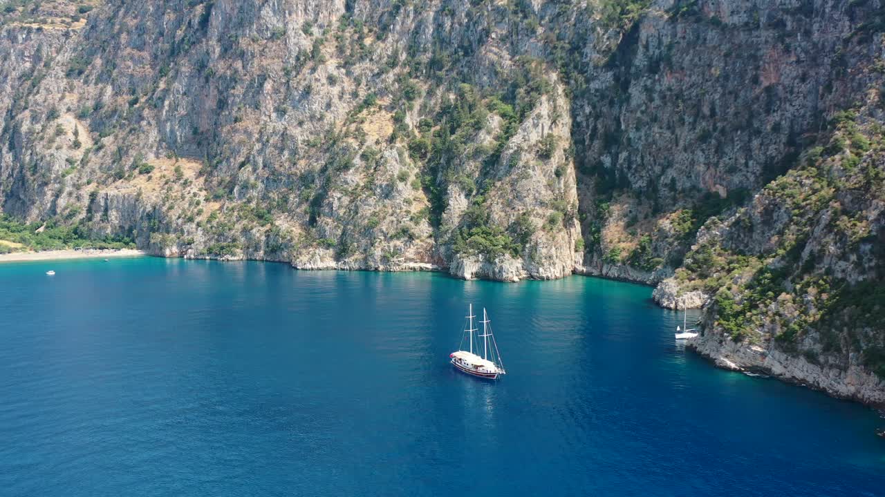 aerial drone circling a large sailboat anchored in the blue ocean of Butterfly Valley panning upwards to the green mountains of Fethiye Turkey on a sunny summer day