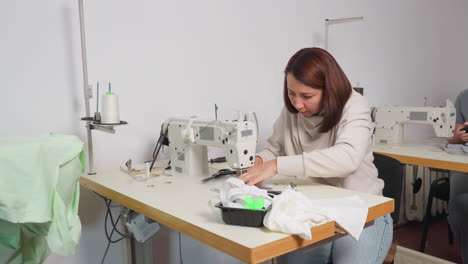 focused tailor works attentively on sewing machine at organized workstation with fabric, scissors, and thread around, while another tailor in background handles fabric, creating busy fashion studio