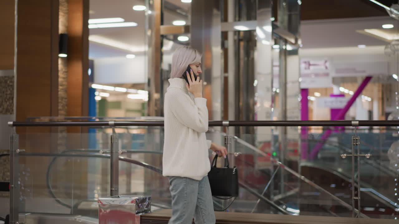 elegant lady walking to bench in mall while talking on phone, placing black purse on bench, wearing white sweater, jeans and boots, glass railing, decorative lights and escalator visible behind