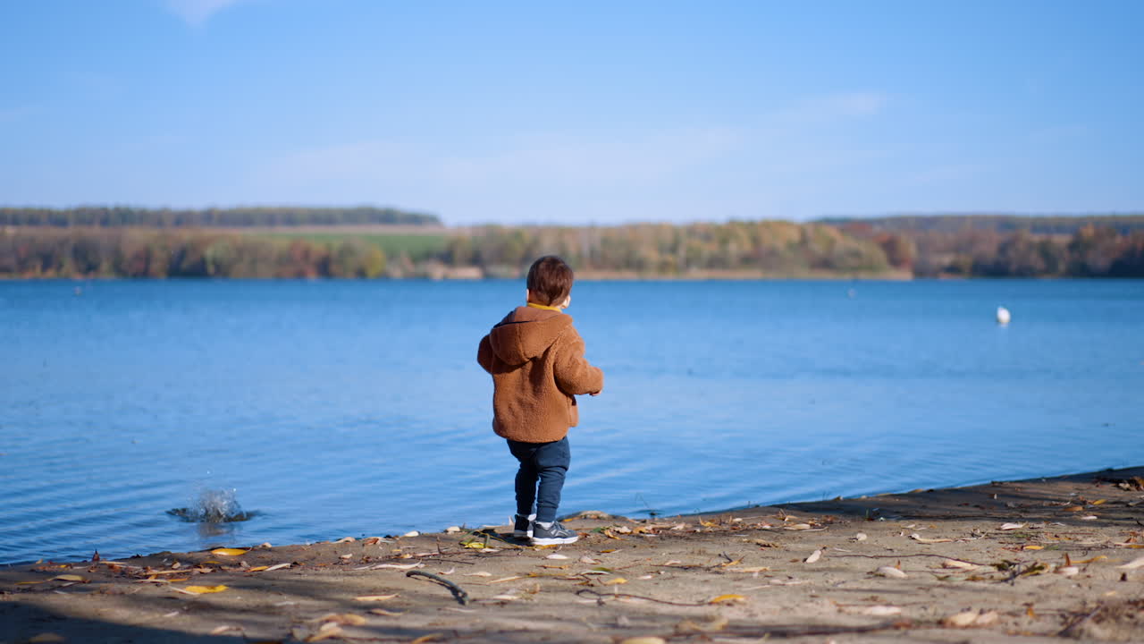 Baby boy spending sunny autumn day at the river. Caucasian toddler throwing the stones into water.