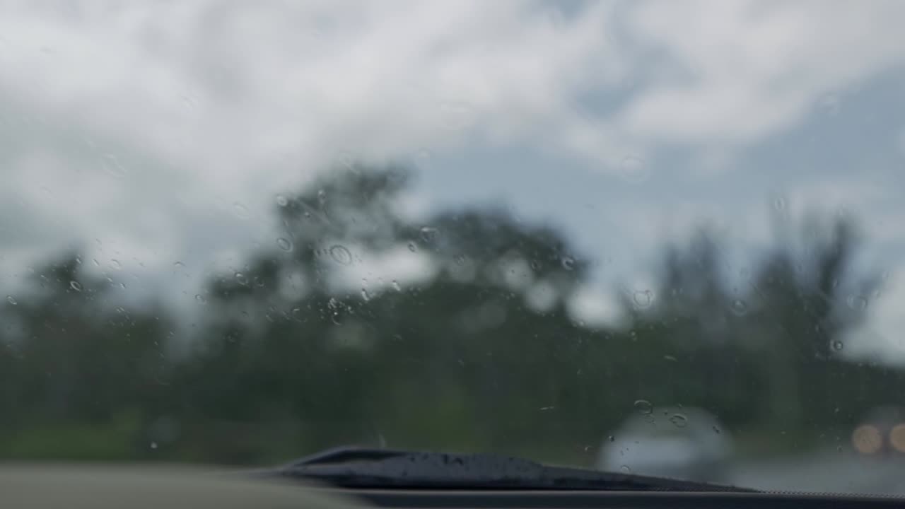 A first-person perspective shot from inside a vehicle, capturing raindrops covering the windshield as the wiper blade sweeps across