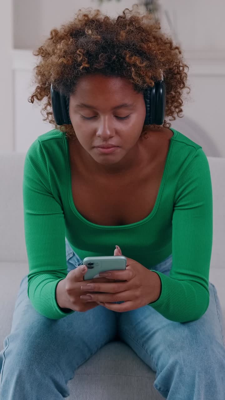 Young woman wearing headphones sits on sofa scrolling smartphone in relaxed home