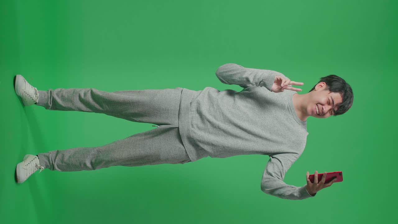 Full Body Of Smiling Asian Man Making Selfie On Mobile Phone While Standing On Green Screen In The Studio