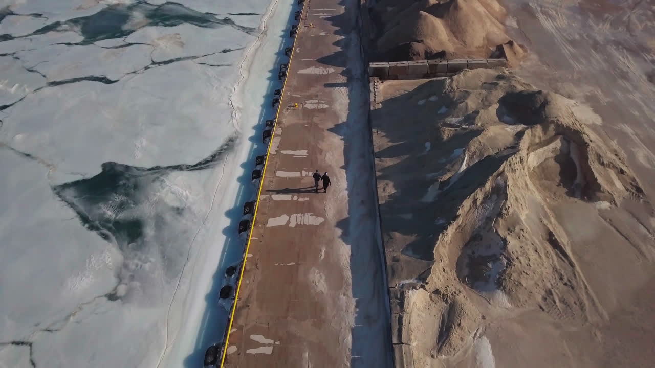 A couple walks down a dock next to a blue frozen icy lake and a stone quarry in winter